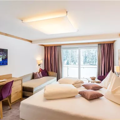 Ein modernes Hotelzimmer mit einem großen Bett, einem Schreibtisch und einem Fernseher. Große Fenster sorgen für viel Tageslicht und einen Blick nach draußen.