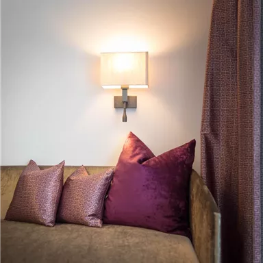 Ein komfortables Sofa mit lila Kissen steht an einer Wand. Eine Wandlampe sorgt für sanftes Licht, ergänzt von einem eleganten Vorhang.