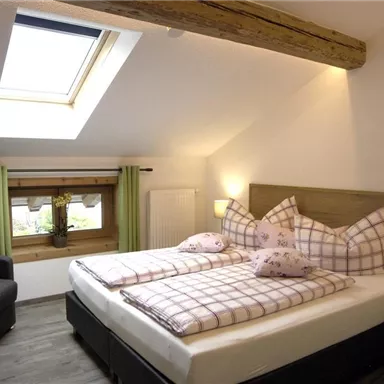 A cozy bedroom with a large window and sloped ceiling. There are two beds with checkered bed covers and a seating area.