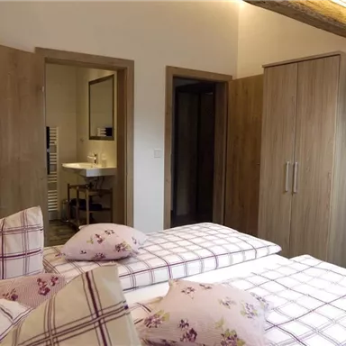 A bright bedroom with two single beds and a modern wardrobe. In the background, a bathroom can be seen that is accessible through a door.