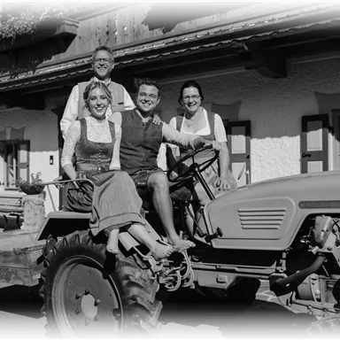 Eine Gruppe von vier Personen posiert auf einem Traktor vor einem traditionellen Bauernhaus. Alle tragen bayerische Trachten und lächeln freundlich in die Kamera.