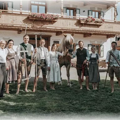 Eine Gruppe von Menschen in traditioneller Tracht steht vor einem ländlichen Gebäude. Sie posieren mit einem Pferd und landwirtschaftlichen Werkzeugen.