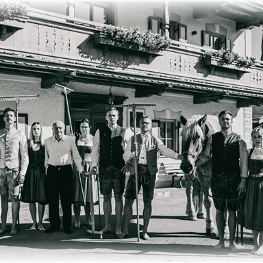 Eine Gruppe von Personen in traditioneller Kleidung steht vor einem ländlichen Gebäude. Die Umgebung zeigt eine rustikale Atmosphäre mit Blumenbalkonen und landwirtschaftlichen Werkzeugen.