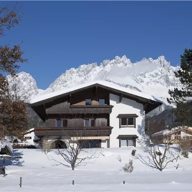 A beautiful alpine house amidst snowy terrain. Impressive mountains and a clear blue sky are visible in the background.