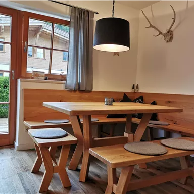 A cozy dining area with a wooden table and benches. The room has large windows overlooking a green terrace.