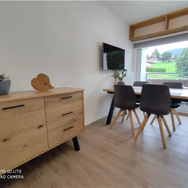 Ein moderner Essbereich mit einem Holztisch und schwarzen Stühlen. Im Hintergrund befindet sich ein Fernseher und große Fenster mit Blick auf die Landschaft.