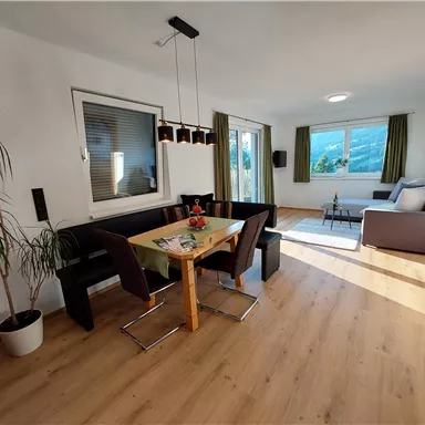 Ein helles und modernes Wohnzimmer mit einem Esstisch und schwarzem Sitzbereich. Große Fenster lassen viel Licht herein und bieten einen Blick nach draußen.