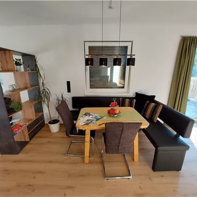 Ein modernes Esszimmer mit einem Holztisch und einem gemütlichen Ecksofa. Der Raum ist hell und freundlich, mit Pflanzen und einer großen Fensterfront.