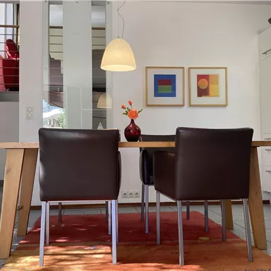 Ein heller Essbereich mit einem großen Holztisch und zwei dunklen Stühlen. Die Küche ist modern und gut ausgestattet, mit bunten Bildern an der Wand.