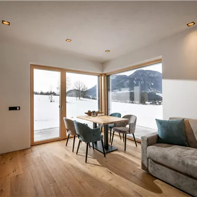 Ein gemütlicher Wohnbereich mit einem Tisch und Stühlen, großes Fenster mit Blick auf die verschneite Landschaft. Das Interieur ist hell und modern gestaltet.