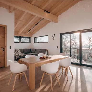 Ein modernes Esszimmer mit einem Holztisch und weißen Stühlen. Große Fenster lassen Tageslicht herein und bieten einen Blick auf die Natur.