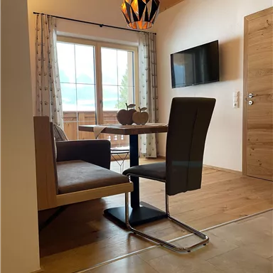 Ein gemütlicher Essbereich mit einem Tisch, einem Stuhl und einer Bank. Große Fenster lassen viel Tageslicht herein und bieten einen schönen Blick nach draußen.