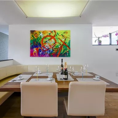 Ein modernes Esszimmer mit einem großen Holztisch und weißen Stühlen. An der Wand hängt ein buntes Kunstwerk, und auf dem Tisch stehen Gläser und eine Flasche Wein.