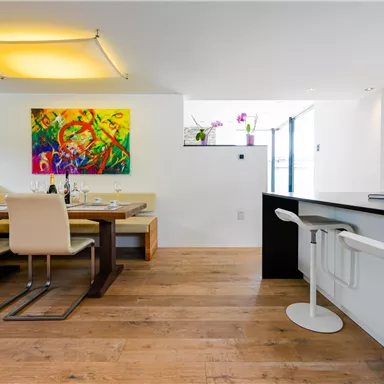 Ein modernes Esszimmer mit einem Holztisch und eleganten Stühlen. An der Wand hängt ein farbenfrohes Gemälde, und große Fenster sorgen für viel Licht.