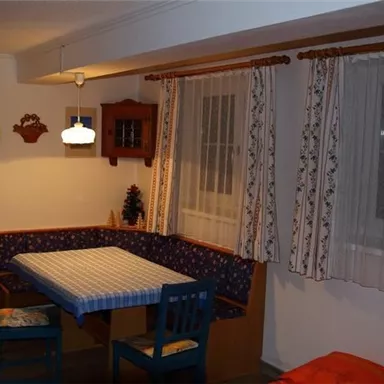 Ein gemütlicher Essplatz mit einem blauen Tisch und einer bankartigen Sitzgelegenheit. Die Wände sind hell gestrichen und es gibt Vorhänge sowie dekorative Elemente.