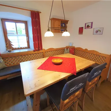 Ein gemütlicher Essbereich mit einem großen Holztablett und einer roten Tischdecke. Die Wände sind freundlich mit Fenstern und dekorativen Elementen geschmückt.