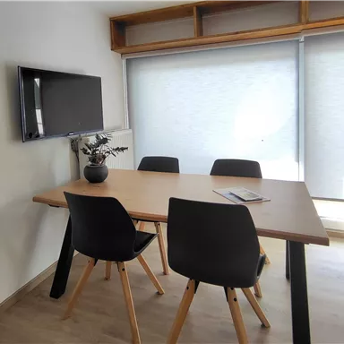 Ein modernes Esszimmer mit einem Holzst am Tisch und vier schwarzen Stühlen. An der Wand befindet sich ein Fernseher und große Fenster mit Jalousien.