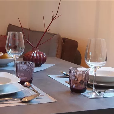 A stylishly set table with white plates, glasses, and candles. In the background, a fruit bowl and a decorative vase are visible.