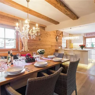 Ein einladender Essbereich mit einem großen Holztisch und geschmackvoller Tischdekoration. Im Hintergrund sieht man eine moderne Küche und große Fenster, die viel Licht hereinfällen.