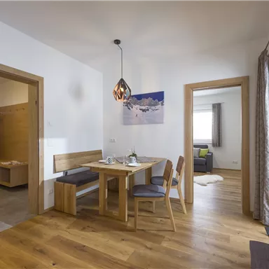 A bright and modern living room with a dining table and chairs. The room has large windows and a cozy atmosphere.