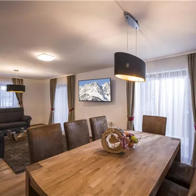 Ein moderner Wohnraum mit einem großen Esstisch und bequemen Stühlen. Im Hintergrund sind ein Sofa und ein Fernseher mit einer Berglandschaft zu sehen.