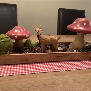 Eine dekorative Tischanordnung mit roten Pilzen, einem Holzreh und grünen Elementen. Die Deko ist auf einem karierten Tischläufer platziert.