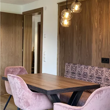 An elegant dining area with a wooden table and pink chairs. The walls are in a warm wood tone, and the lighting creates a cozy atmosphere.