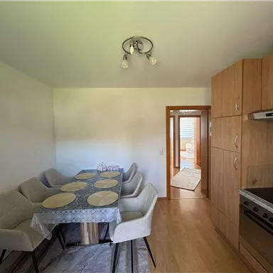 Eine moderne Küche mit einem großen Esstisch und beigen Stühlen. Im Hintergrund befindet sich eine Tür, die zu einem weiteren Raum führt.