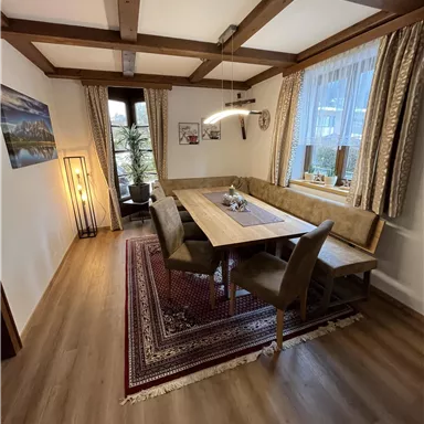 Ein gemütlicher Essbereich mit einem großen Holztisch und gepolsterten Stühlen. Große Fenster lassen viel Licht herein und eine schöne Teppich unterstreicht die warme Atmosphäre.