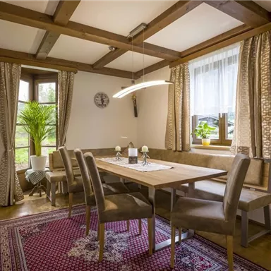 Ein einladender Essbereich mit einem Holztisch und komfortablen Stühlen. Große Fenster lassen viel Licht herein und eine Pflanze sorgt für eine freundliche Atmosphäre.