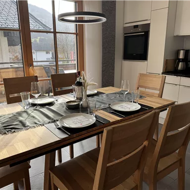 An inviting dining area with wooden chairs and a large table. The table is set with dishes and glasses, and large windows provide a view of the surroundings.