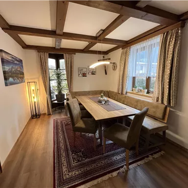 Ein gemütlicher Essbereich mit einem großen Holztisch und bequemen Stühlen. Die Wände sind mit Bildern und Vorhängen dekoriert, und das Licht ist warm und einladend.