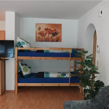 Ein einfaches Zimmer mit einem Etagenbett und einer gemütlichen Sitzecke. An der Wand hängt ein Kunstwerk mit Blumenmotiven.