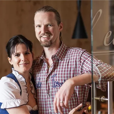 Ein Paar lächelt in traditioneller Kleidung. Sie stehen in einem gemütlichen Raum mit Holzdetails.