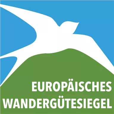 Ein Logo des Europäischen Wandergütesiegels zeigt einen Vogel über einem grünen Hügel vor einem blauen Himmel. Darunter steht in großen Buchstaben „EUROPÄISCHES WANDERGÜTESIEGEL“.