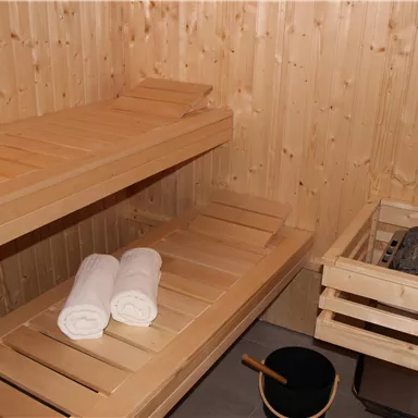 Eine gemütliche Sauna aus Holz mit zwei Bänken und sauberen Handtüchern. Neben der Saunabank befinden sich ein Saunaofen und ein Eimer.
