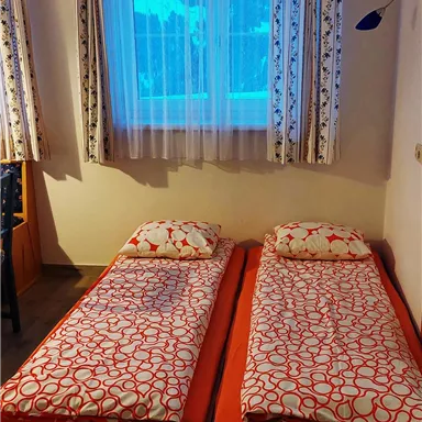 Ein einfaches Schlafzimmer mit zwei Einzelbetten und bunten Bettdecken. Das Fenster lässt viel Tageslicht herein und zeigt eine angenehme Aussicht.