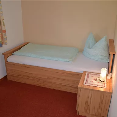 Ein einfaches Schlafzimmer mit einem Einzelbett und einem Nachttisch. Die Wände sind hell gestrichen, und es gibt ein Fenster mit Vorhängen.