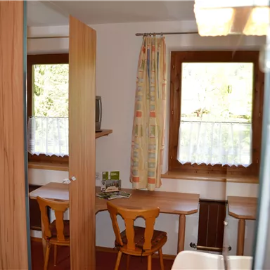 Ein gemütlicher Raum mit zwei Tischen und Stühlen. Neben einem Fenster mit Vorhängen sind einige Pflanzen und ein Fernseher sichtbar.