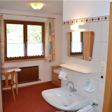 Ein einfaches Badezimmer mit einem Waschbecken, einem Spiegel und einer Duschecke. Im Hintergrund steht ein kleiner Tisch mit Stühlen und ein Fenster neben dem Eingang.