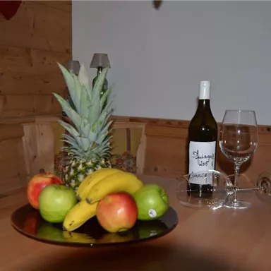 A table with a fruit bowl displaying pineapples, apples, pears, and bananas. Next to it is a bottle of white wine and glasses.