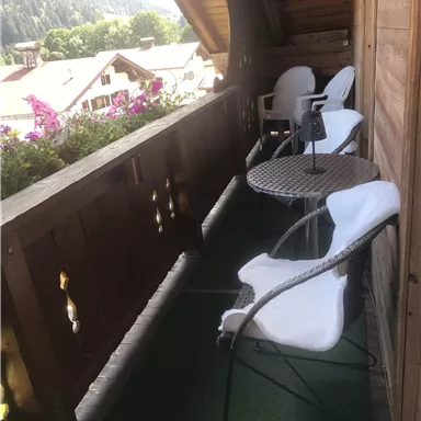 A cozy balcony with white chairs and a small table. In the background, green mountains and colorful flowers can be seen.