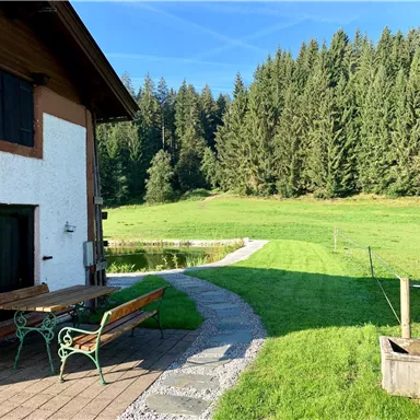 Ein charmantes Haus mit einem bepflanzten Garten und einem Weg, der durch eine Wiese führt. Im Hintergrund sind dichte, grüne Wälder zu sehen.