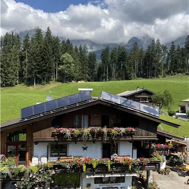 Ein schönes Holzhaus mit Blumenschmuck und Solarpanelen. Im Hintergrund sind grüne Wiesen und bewaldete Berge zu sehen.