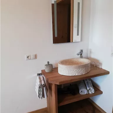 A modern bathroom with a wooden vanity and a round wash basin. In the background, there is a mirror as well as towels and glasses on the shelf.