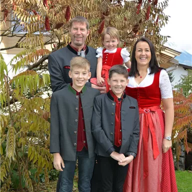 Eine Familie in traditioneller Kleidung steht vor einem Baum mit bunten Blättern. Die Kinder lächeln und die Erwachsenen haben festliche Trachten an.