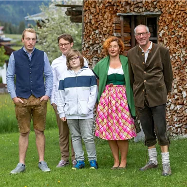 Eine Gruppe von fünf Personen steht im Freien vor einem Holzstapel. Sie sind in traditioellen Kleidern gekleidet und lächeln freundlich in die Kamera.
