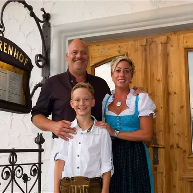 A friendly couple and a boy are standing in front of a wooden gate with a menu view. The woman is wearing a traditional alpine dress, while the boy is wearing a white blouse and trousers.