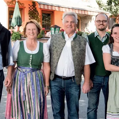 Eine Gruppe von fünf Personen in traditioneller Kleidung steht zusammen. Im Hintergrund sind ein paar grüne Pflanzen und ein Gebäude sichtbar.