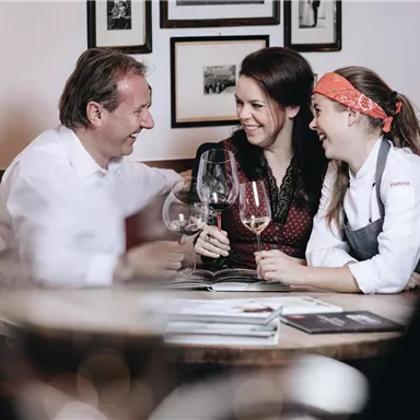 Zwei Frauen und ein Mann sitzen an einem Tisch, lachen und stoßen mit Weingläsern an. Im Hintergrund sind Bilder an der Wand zu sehen.